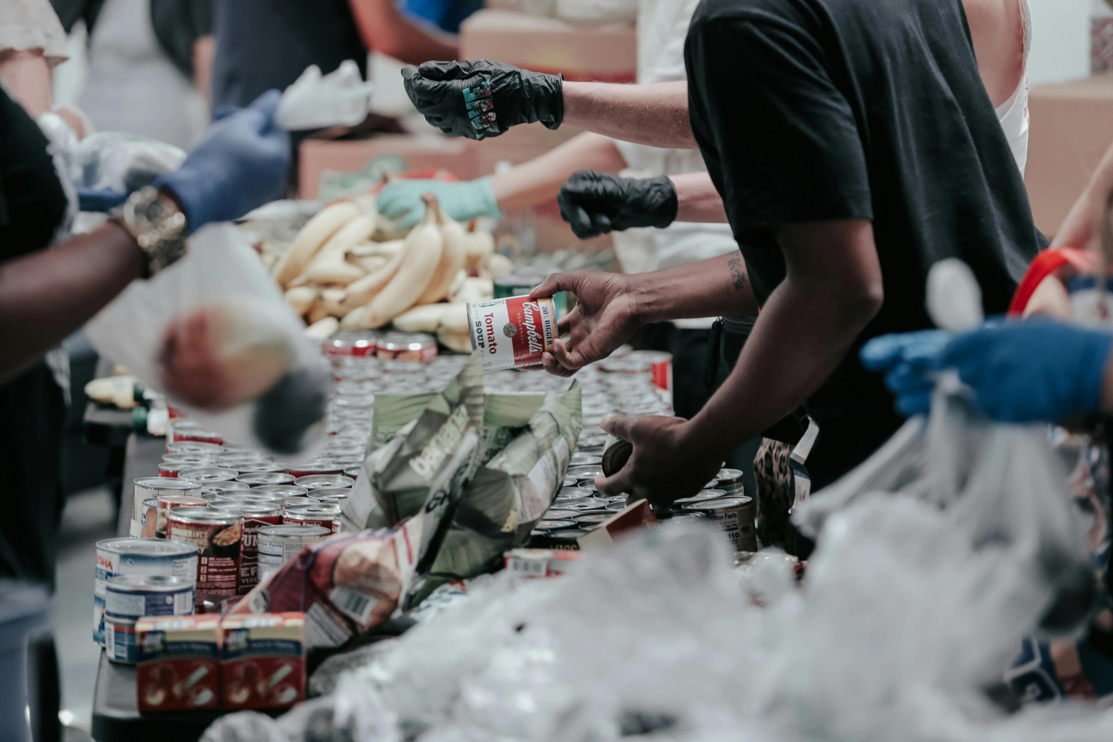 Shows a food drive with a table full of different types of food and volunteers giving out donations